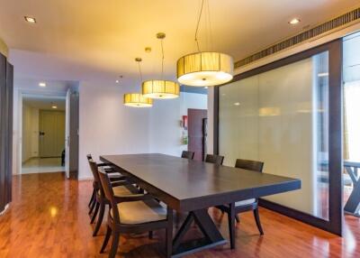 A modern dining room with wooden flooring and a long table with six chairs, pendant lighting, and glass partition.