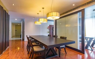 A modern dining room with wooden flooring and a long table with six chairs, pendant lighting, and glass partition.