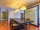 A modern dining room with wooden flooring and a long table with six chairs, pendant lighting, and glass partition.