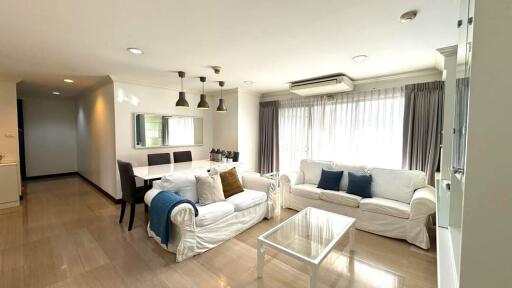 A spacious and well-lit living room with large windows, white sofas, a dining table, and modern lighting fixtures.