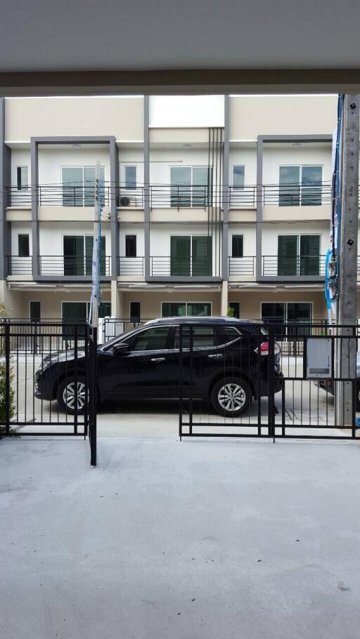 View of a townhouse building with multiple floors and balconies, seen from a driveway with a parked black car in front