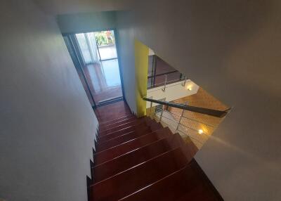 View of a wooden staircase with a metal railing, leading to a sunlit room with large windows.