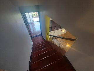 View of a wooden staircase with a metal railing, leading to a sunlit room with large windows.