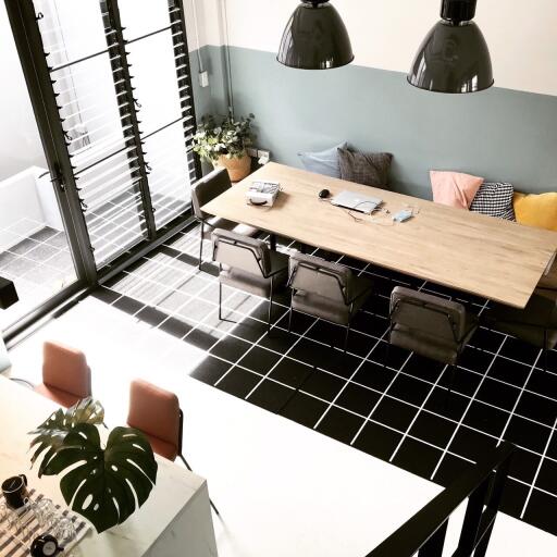 Modern dining room with large wooden table, four chairs, pendant lights, and a mix of decorative pillows on sofa with access to balcony.