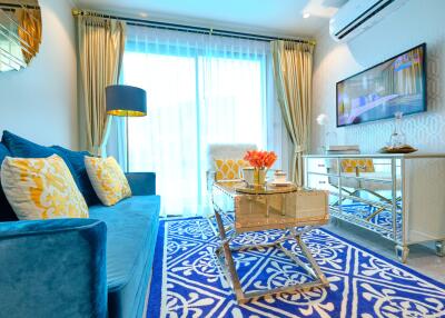 A well-furnished living room with a blue sofa, yellow cushions, a decorative rug, a glass coffee table, a TV, and elegant curtains.