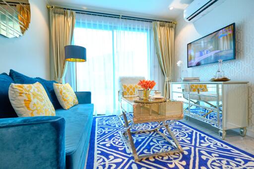 A well-furnished living room with a blue sofa, yellow cushions, a decorative rug, a glass coffee table, a TV, and elegant curtains.