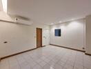 Spacious empty living room with white tiles, wooden accents, window, and air conditioning unit