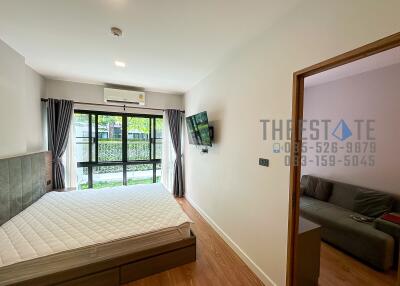 Well-lit bedroom with bed, wall-mounted TV, large window and a view of greenery.