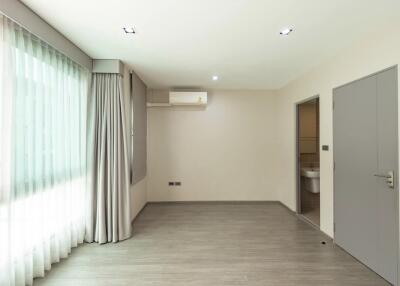 Spacious bedroom with large window, grey curtains, wooden floor, air conditioning, and adjacent bathroom.