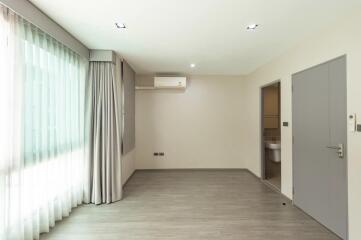 Spacious bedroom with large window, grey curtains, wooden floor, air conditioning, and adjacent bathroom.