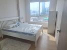 Bright bedroom with a large window, a bed with white frame, bedside tables, and a city view