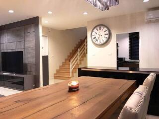 Modern living room with wooden dining table, wall clock, staircase, and wall-mounted TV