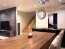 Modern living room with wooden dining table, wall clock, staircase, and wall-mounted TV