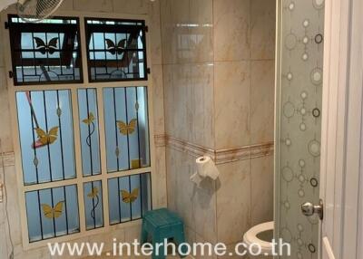 Bathroom with tiled walls and floor, featuring a glass shower door, window with decorative grills, toilet, and small fan.