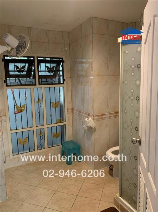 Bathroom with tiled walls and floor, featuring a glass shower door, window with decorative grills, toilet, and small fan.