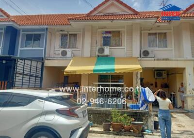 Front view of a townhouse with a yellow and green striped awning, white car parked in front, and a person hanging clothes.