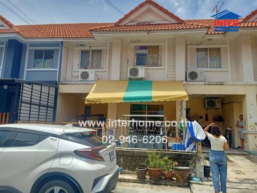 Front view of a townhouse with a yellow and green striped awning, white car parked in front, and a person hanging clothes.