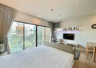 A bright studio apartment with large windows, balcony, bed, wall-mounted TV, small dining table, and modern decor.