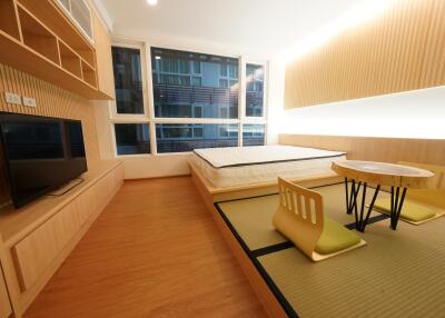 Modern bedroom with a minimalist design featuring a large window, wooden flooring, a bed, a TV on a wooden console, built-in shelving, and a unique tatami area with a small table and chairs.