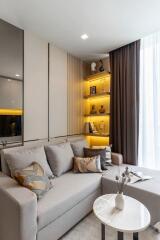 Modern living room with a grey sofa, decorative pillows, a round white marble coffee table, built-in shelves with warm lighting, and a large window with curtains.