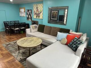 Living room with white sectional sofa, decorative pillows, artworks on teal walls, parquet flooring, and dining area