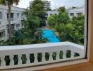 View from a window overlooking a swimming pool and lush greenery in between multiple white buildings