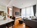 Modern living room with a large window, comfortable gray sofa, wall-mounted TV, wooden floor, and dining area.