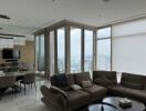 Modern living room with large windows, a grey sectional sofa, dining area, and city skyline view