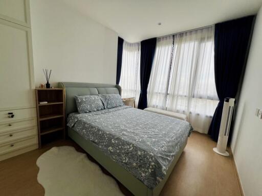 A tidy bedroom with a bed, nightstand and large windows with curtains