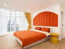 Bright modern bedroom with large windows, red and white bedding, yellow nightstand, and wooden flooring.