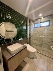 Modern bathroom with green tiled accent wall, round mirror, glass-enclosed shower, and contemporary fixtures.