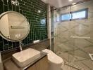 Modern bathroom with green tiled accent wall, round mirror, glass-enclosed shower, and contemporary fixtures.