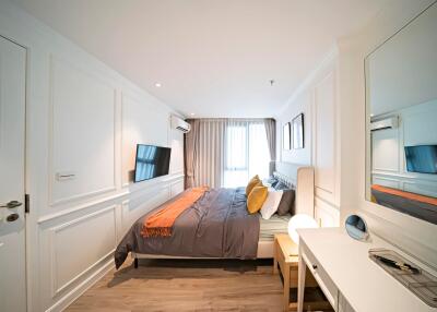 Modern bedroom with grey and orange bedding, wall-mounted TV, desk with mirror, and large window