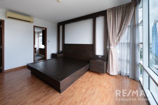 Spacious bedroom with a mounted air conditioner, large window with curtains, wooden flooring, and a dark wood bed frame.