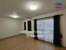 Spacious living room with wooden floor, large windows covered with black curtains and an air conditioning unit.