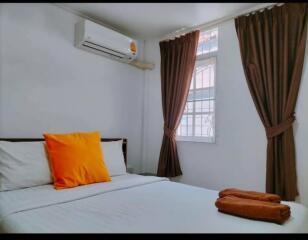 A bedroom with a neatly made bed, a window with brown curtains, and an air conditioning unit.