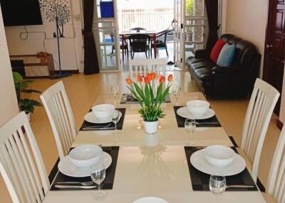 Modern dining room with a glass table set for six, featuring a centerpiece of tulips, open to a living area with a TV and sofa and leading to a patio with large windows.