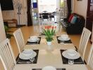 Modern dining room with a glass table set for six, featuring a centerpiece of tulips, open to a living area with a TV and sofa and leading to a patio with large windows.