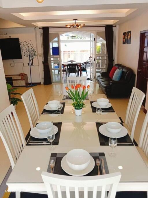 Modern dining room with a glass table set for six, featuring a centerpiece of tulips, open to a living area with a TV and sofa and leading to a patio with large windows.