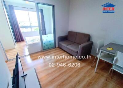 Modern living area with wooden floor, sofa, TV, dining table, and bedroom visible through sliding glass doors