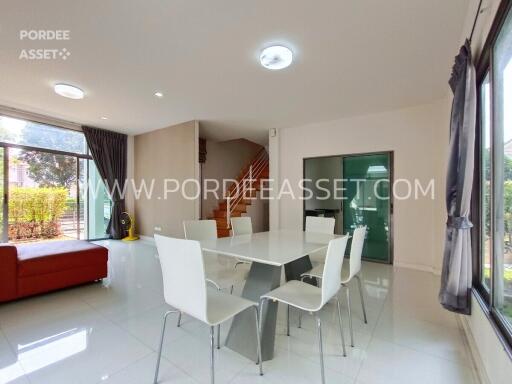 Modern dining area with white table and chairs, adjacent to living room with staircase and large windows.