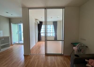 Modern living room with an adjacent bedroom separated by a glass partition, both featuring wooden floors.