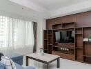 Modern living room with built-in wooden shelves, a wall-mounted TV, large windows with curtains, and a coffee table with a flower vase.