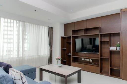 Modern living room with built-in wooden shelves, a wall-mounted TV, large windows with curtains, and a coffee table with a flower vase.