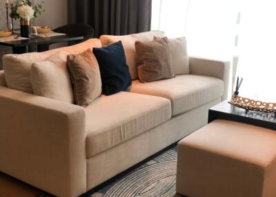 A cozy living room with a beige sofa and matching ottoman, accented with pillows and a nearby dining setup.