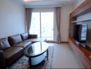 Modern living room with a large brown leather sectional sofa, a glass coffee table, a flat-screen TV on a wooden media console, and large sliding glass doors leading to a balcony.