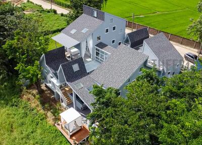 Aerial view of a modern house with multiple small buildings and a grey shingled roof surrounded by greenery