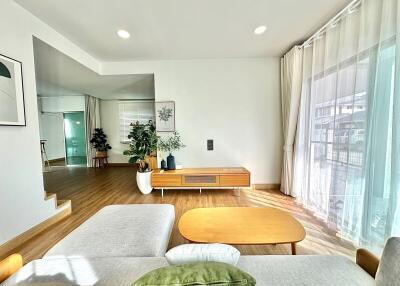 Modern living room with wooden flooring and furniture, large window with curtains, and indoor plants