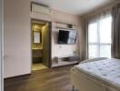 Bedroom with wooden flooring, large window, mounted TV, and attached bathroom