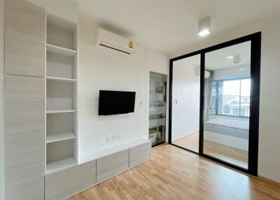 Modern living area with built-in shelves, wall-mounted TV, air conditioning, and view of the bedroom.